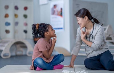 A female therapist sits beside her young patient on the floor as they work on some speech therapy exercises.  They are each dressed casually and have their hands on their chins as they work through phonetical sounds.   Small white letter tiles can be seen laying out in front of them from a previous exercise.